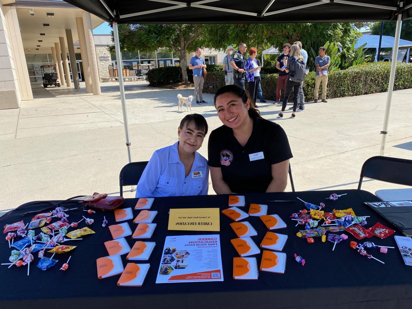 Career Education Table at Job Fair.  Two Classified Professionals sitting and smiling at table.